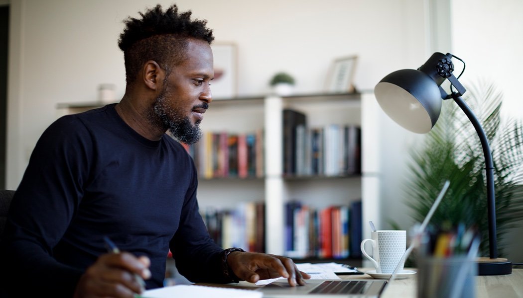 A Black man sits in front of his computer while taking notes on his laptop.