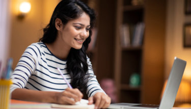 A woman writes down notes on her notepad while smiling at her computer.