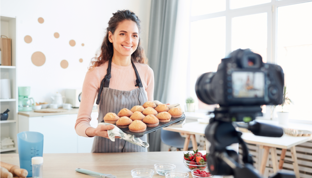 A woman holds muffins on a baking sheet in front of the camera recording it.