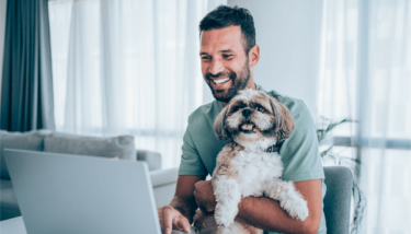 A man holds a dog while smiling at his computer when looking for membership site ideas.