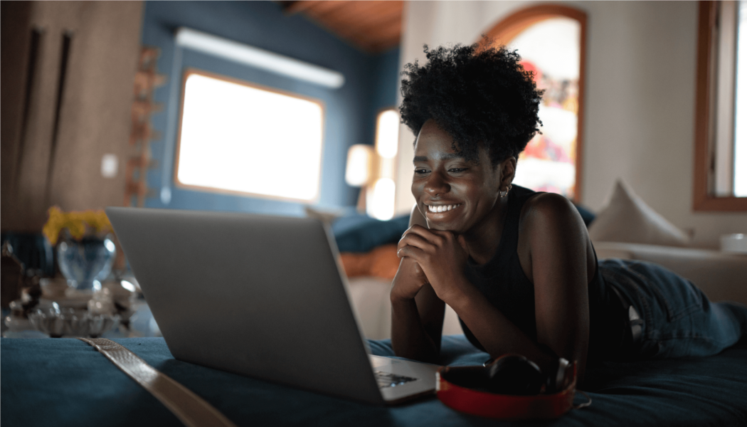 A woman lies on her bed while smiling at her computer screen.