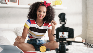 A woman sits on her bed and smiles at the camera while recording a video.