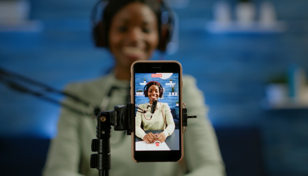 A content creator smiles while live streaming, captured through a smartphone mounted on a tripod. The screen displays the creator wearing headphones, sitting at a desk with a microphone, engaging with her audience on a live streaming app. The background is blurred, focusing attention on the vibrant interaction depicted on the phone's screen.