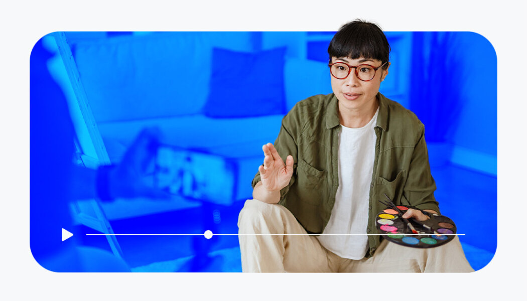 Woman speaking to a camera while holding a paint palette and brushes, behind a stylized blue background.