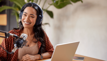 Content creator smiling while sitting at a desk with her laptop, microphone, and headphones.