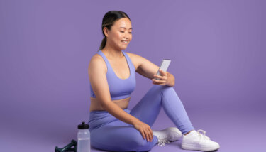 A fitness creator taking a break during her workout session, smiling while browsing through a fitness app on her smartphone, with a water bottle and dumbbells placed beside her on a purple background.