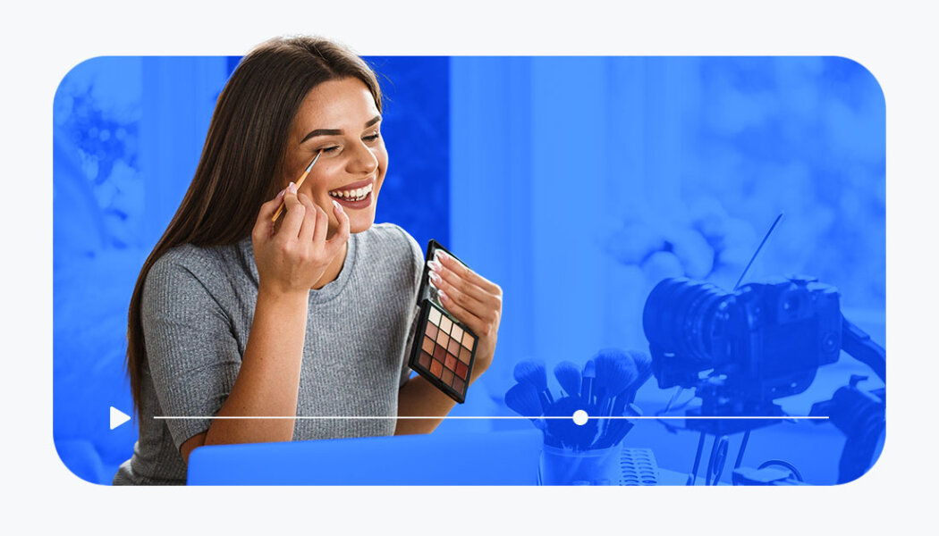 Woman filming an eyeshadow tutorial with a camera in front of a desk with a blue filtered background.